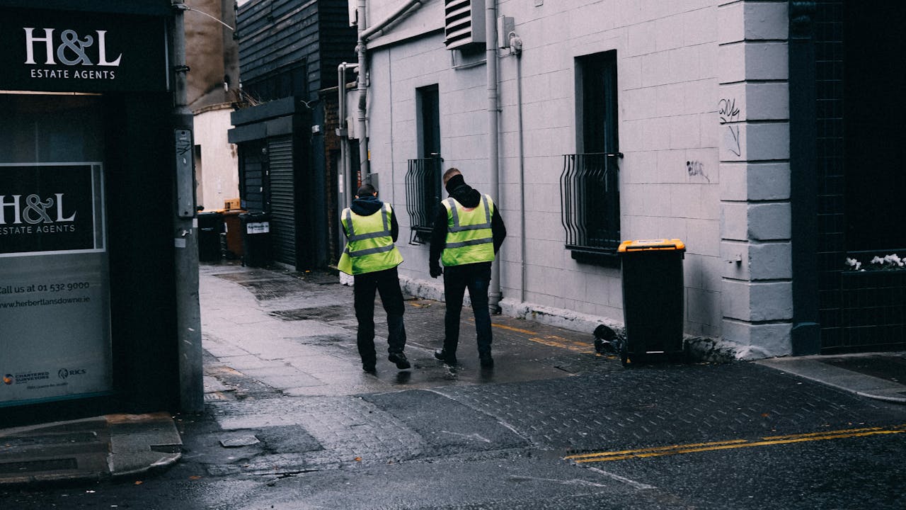 Two safety-vested men walk in a rainy urban alley, next to estate agency.