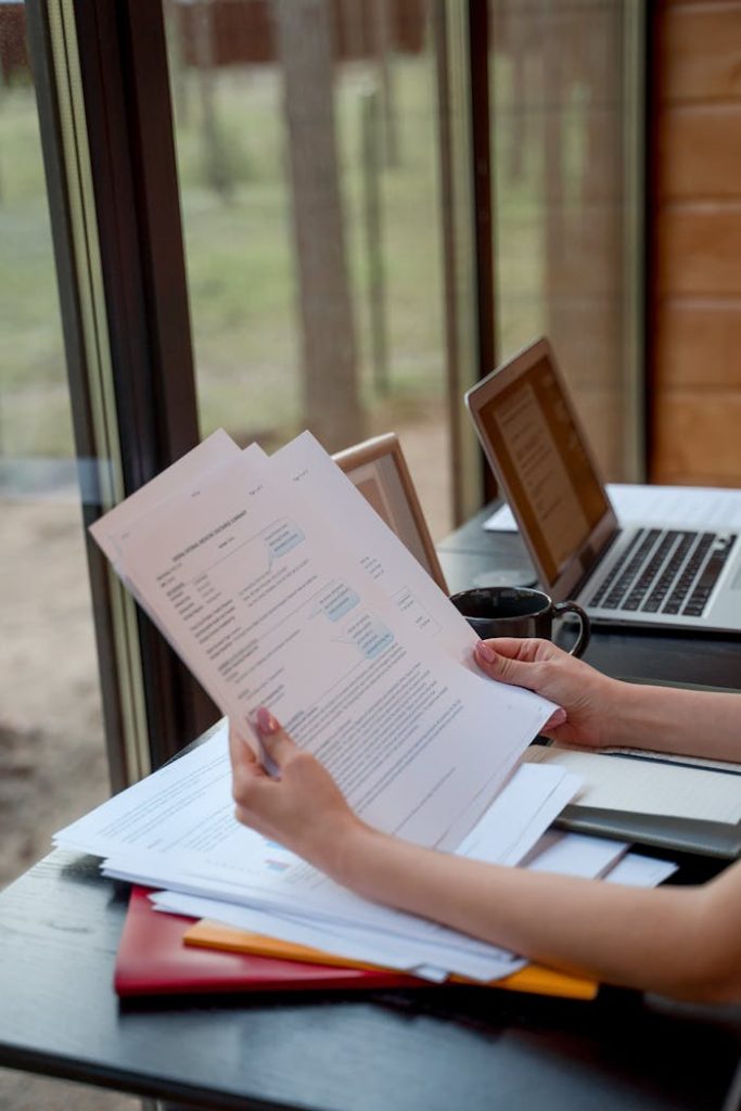 Person holding and reviewing documents near window with laptops on a desk.